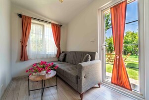 Ένα άνετο δωμάτιο με έναν καναπέ, ένα μικρό τραπέζι στολισμένο με λουλούδια και μεγάλα παράθυρα με κουρτίνες.