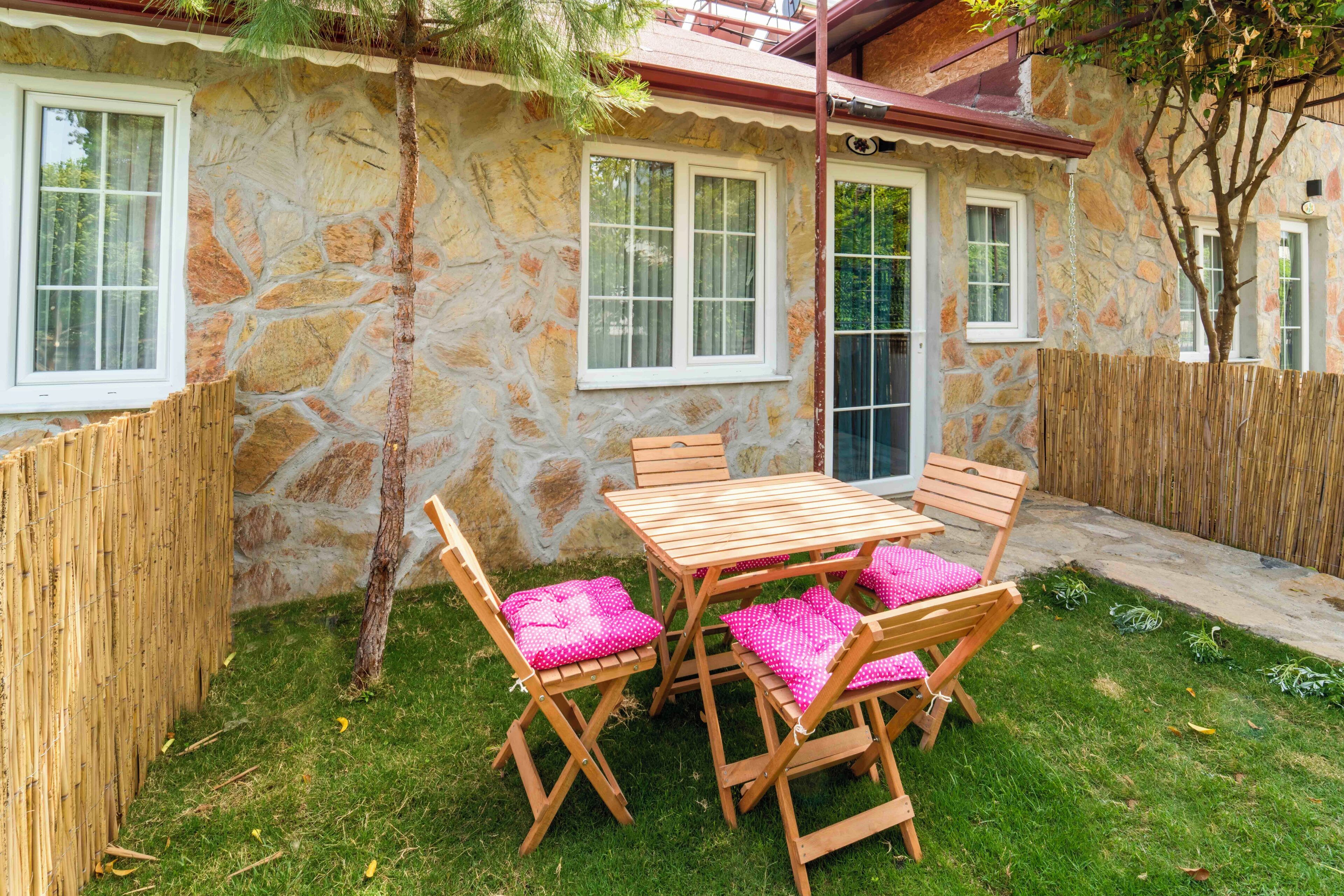 Een stenen huis met een houten tafel en stoelen, een bamboenschutting en een kleine boom.