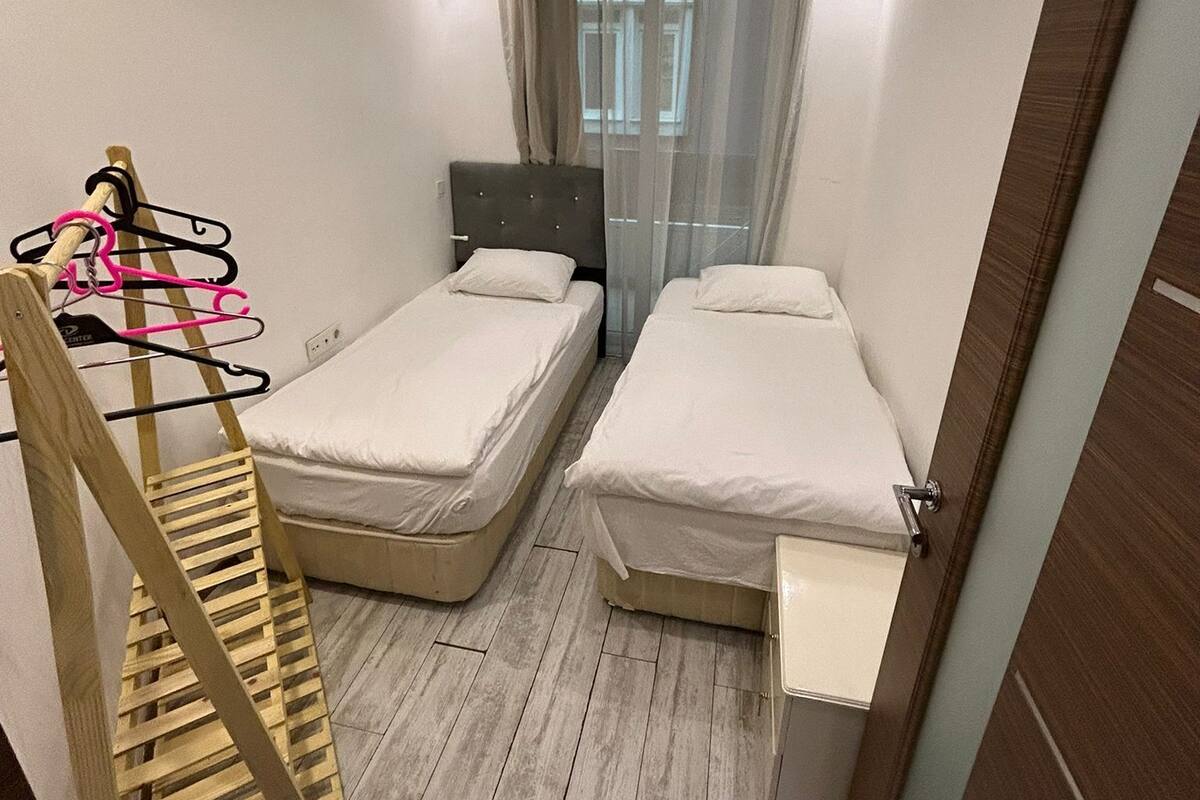 Ein kleines, helles Zimmer mit zwei Einzelbetten, einer Holzleiter, einem kleinen Schreibtisch und einem Fenster mit durchsichtigen Vorhängen.