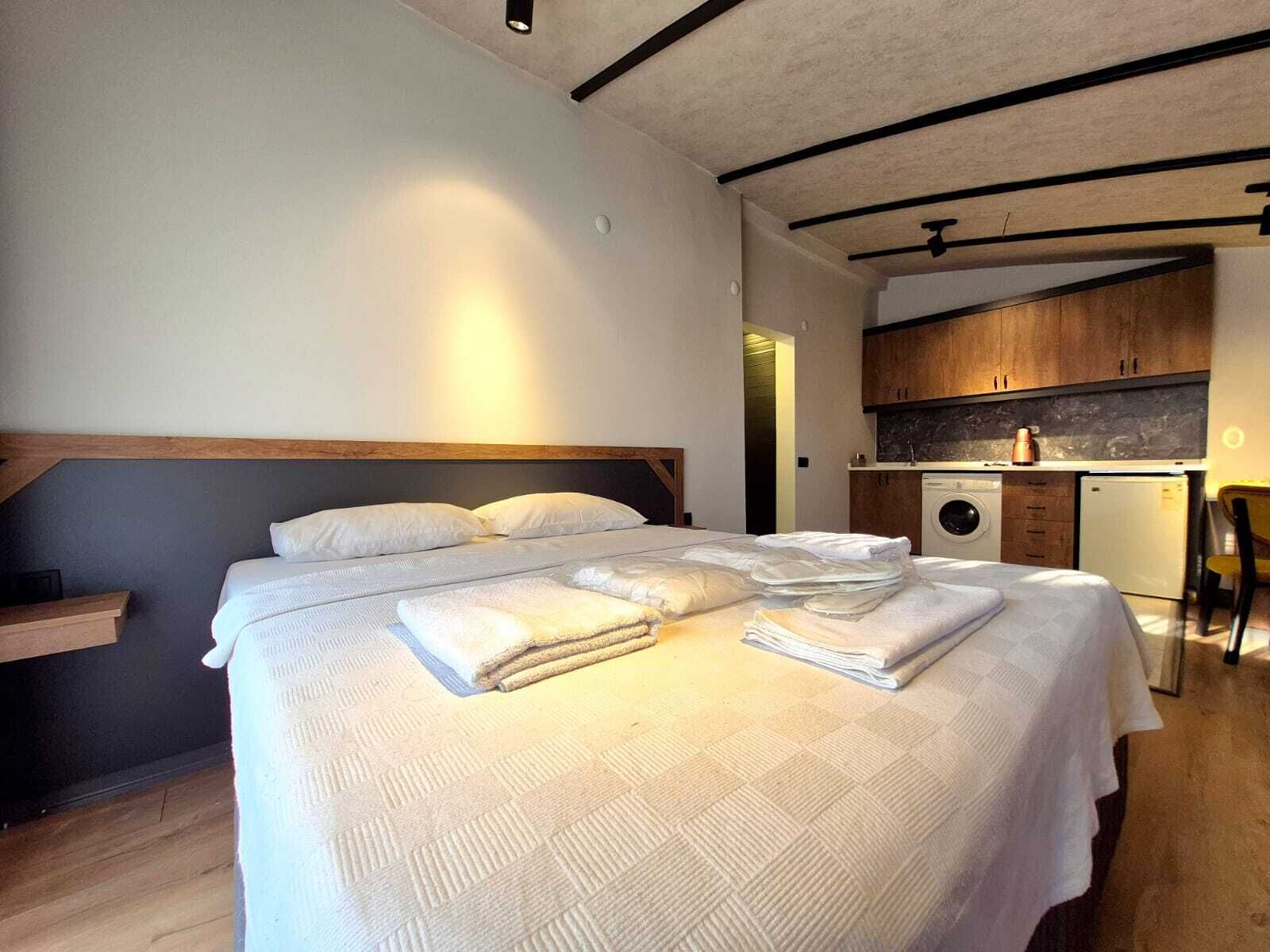 A bedroom with a bed, a bedside table, a washing machine, and a small kitchen area.
