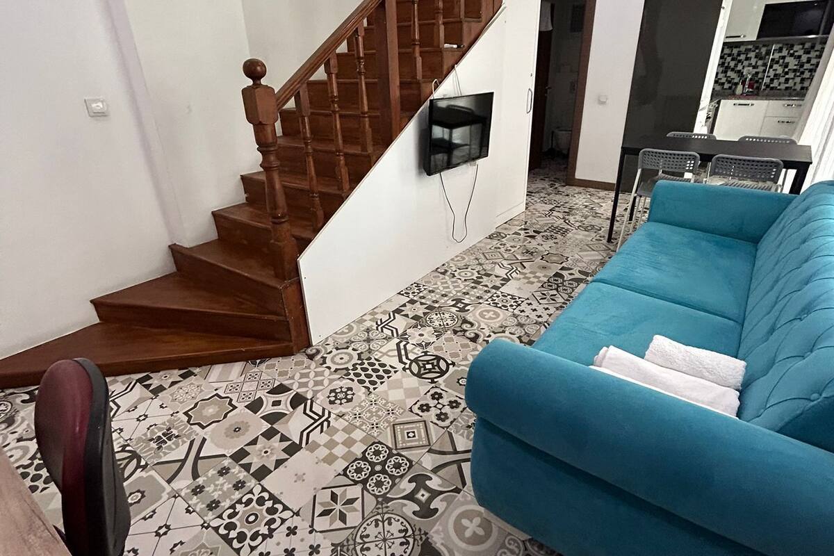 Ein Wohnzimmer mit einem gemusterten Boden, einem blauen Sofa, einer Holztreppe und einem an der Wand montierten Fernseher.