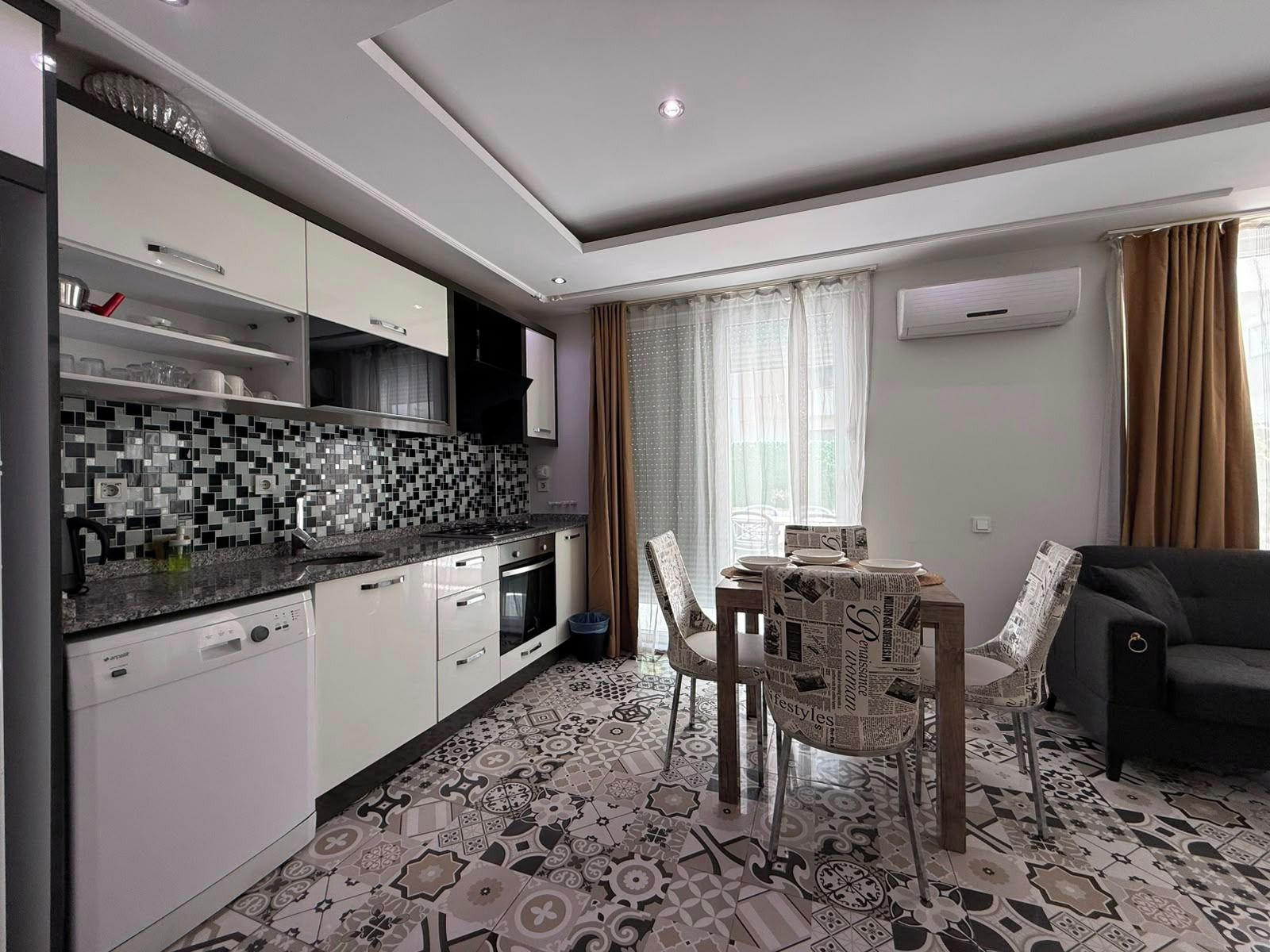 Une cuisine moderne avec des meubles blancs, un dosseret carrelé en noir et blanc, et un coin repas avec une table dressée pour deux personnes.