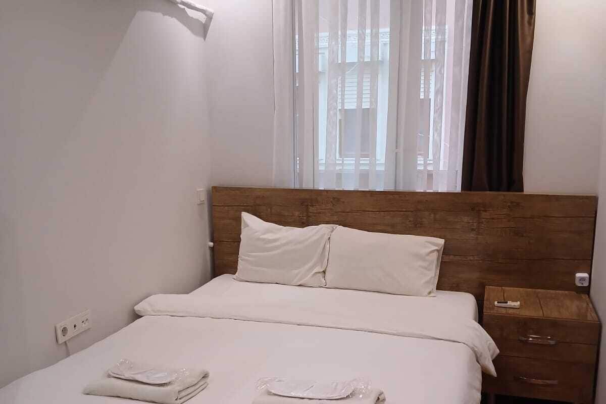Ein ordentlich bezogenes Bett mit weißen Leinen, ein Holzkopfstück, ein Nachttisch mit Fernbedienung und ein Fenster mit durchsichtigen Vorhängen und einem braunen Vorhang.