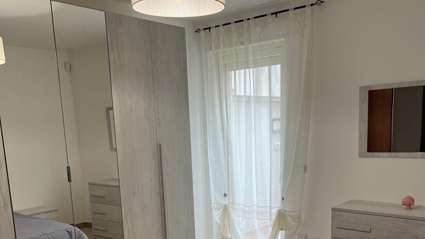 Ein Schlafzimmer mit einem großen Bett, einem Kleiderschrank, einem Spiegel und einem Fenster mit durchsichtigen Vorhängen.