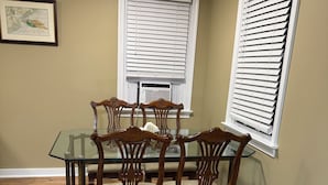Un comedor con una mesa de vidrio y sillas de madera, dos ventanas con persianas y un cuadro en la pared.