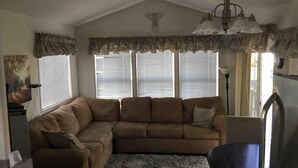 Una sala de estar con un sofá modular, una mesita y un sillón. Hay tres ventanas con persianas, una lámpara de techo y una lámpara de pared.