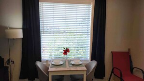 Een eetgedeelte met een tafel gedekt voor twee personen, een raam met jaloezieën, een lamp en een rode stoel.