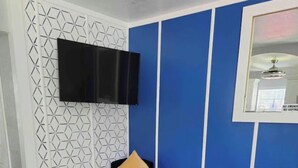 Een moderne woonkamer met een wand in geometrisch patroon, een flatscreen televisie boven een donkerblauwe fauteuil, een klein houten krukje en een grijze poef.
