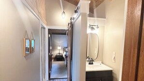 Un pasillo con suelo de madera, pared blanca y techo de madera. Hay un baño con lavamanos y espejo.