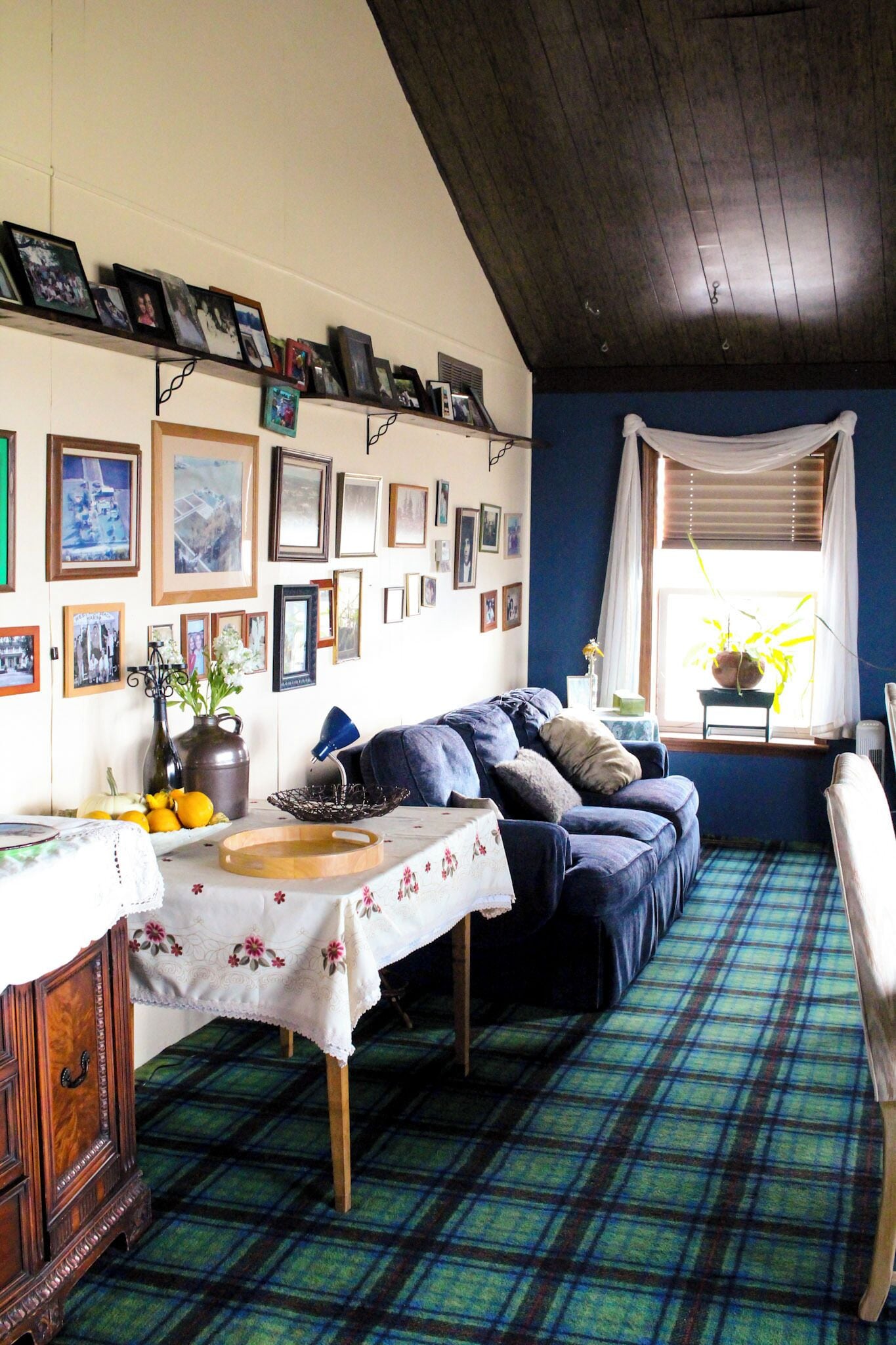 Una habitación acogedora con alfombra escocesa, un sofá, una mesa con mantel floreado y una ventana con cortinas.