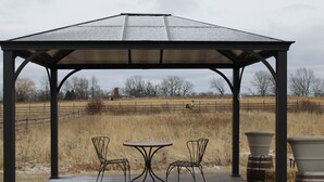 A gazebo with a metal roof, two metal chairs, and a table on a wooden deck.