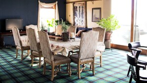 A dining area with a plaid carpet, wooden chairs, a table with a floral tablecloth, potted plants, and a television on a stand.