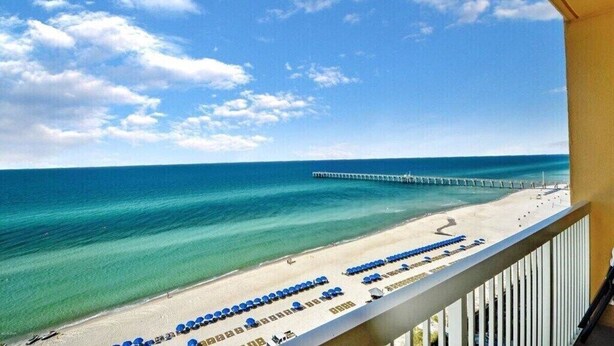 Vista de una playa con un muelle que se adentra en el océano, y una fila de sillas azules a lo largo de la orilla.