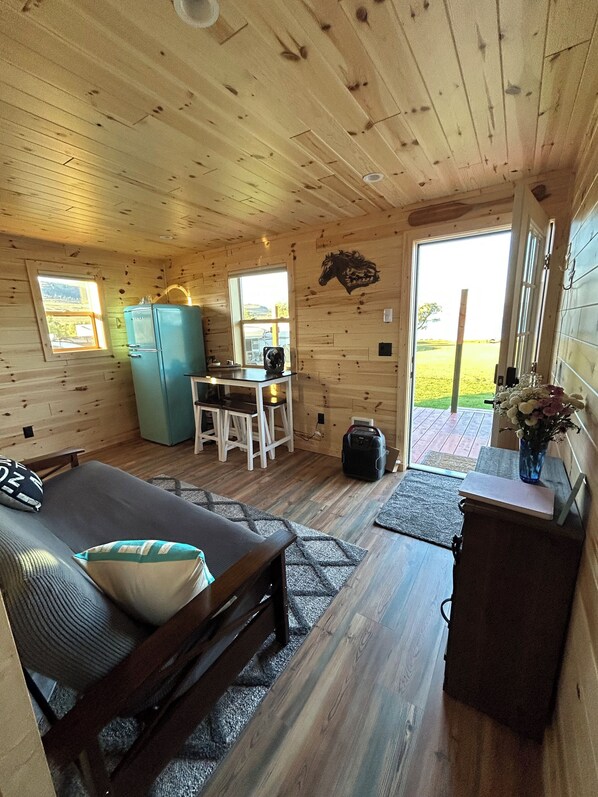 A cozy cabin interior with a bed, a dining table, a refrigerator, and a window overlooking a grassy area.