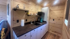 Cozinha compacta com paredes de madeira, bancada preta, armários brancos, pia e cafeteira.