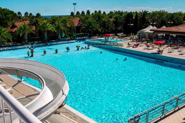 A large outdoor swimming pool with a water slide, surrounded by palm trees and lounge chairs.