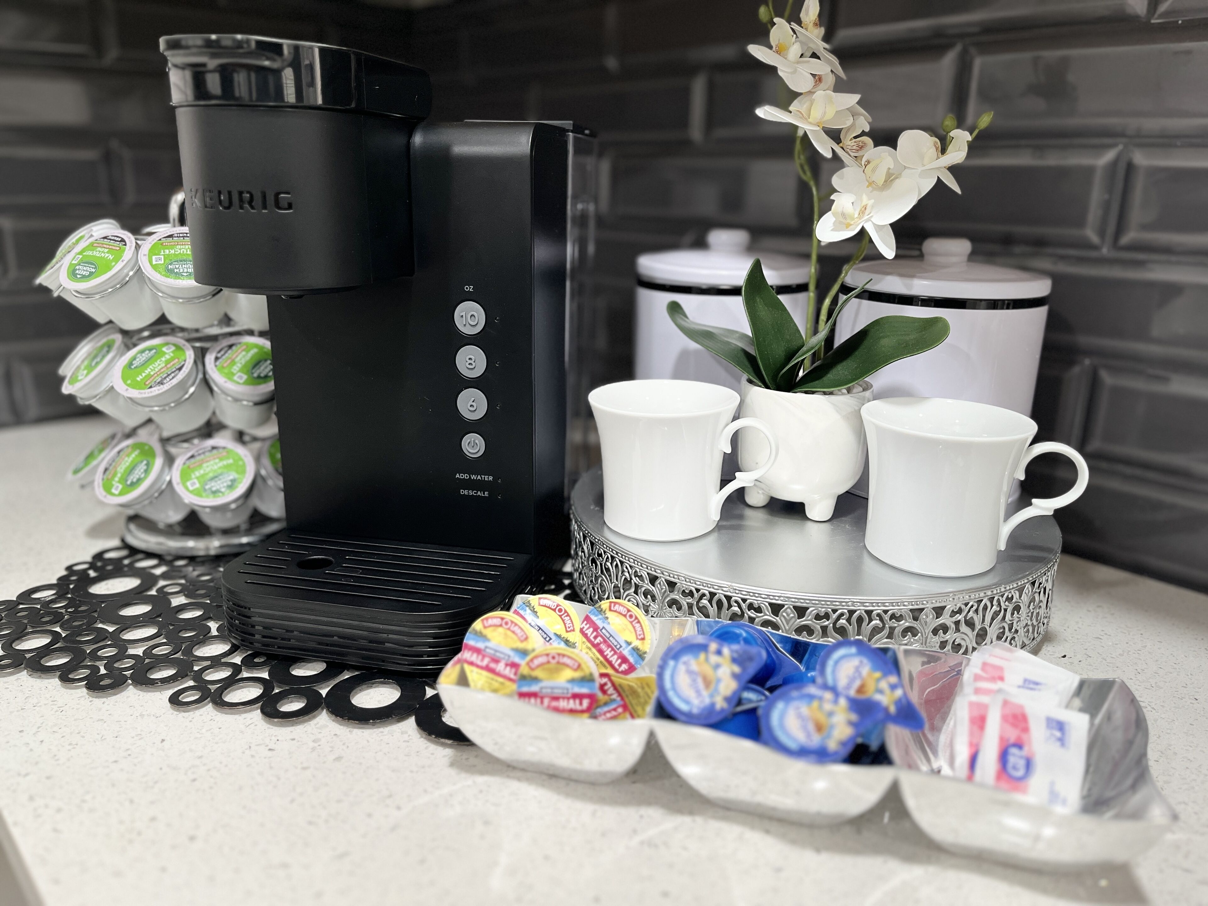 A coffee maker, coffee pods, a plant, and two white cups on a countertop.
