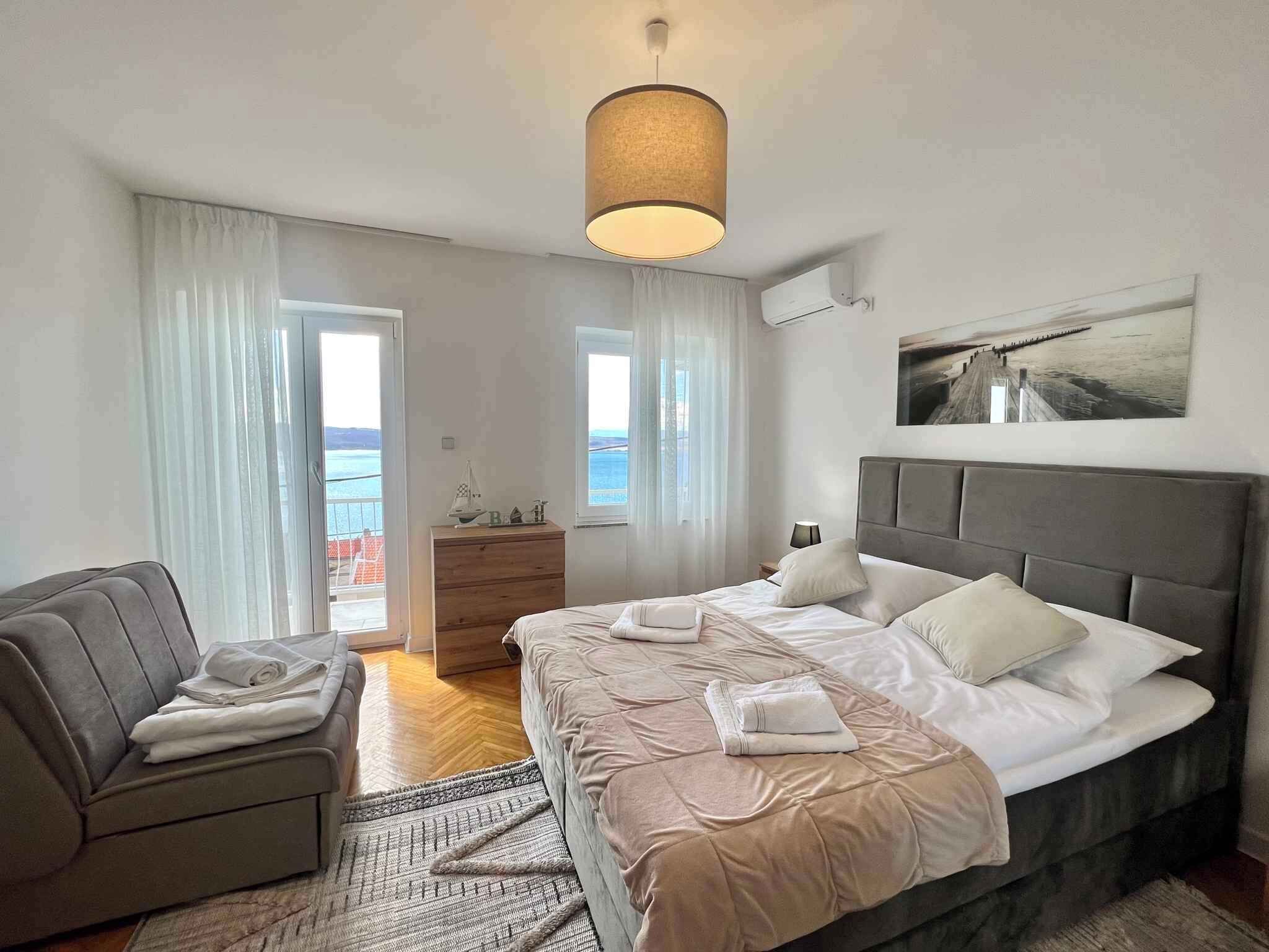 Une chambre à coucher moderne avec un grand lit, un canapé gris, une commode en bois et une vue sur la mer par les fenêtres.