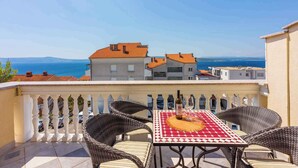 Un balcon agrémenté de fauteuils en osier et d’une table préparée pour deux personnes, d’où l’on peut admirer une ville côtière avec ses bâtiments et la mer.