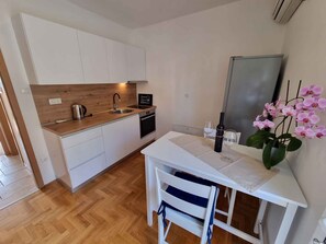 Une cuisine compacte avec des meubles blancs, un plan de travail en bois, un évier et un petit four. On y trouve une table blanche ornée d’un vase contenant des orchidées roses et une bouteille de vin.