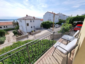 A balcony with a chair, a view of a street with parked cars, and buildings in the background.