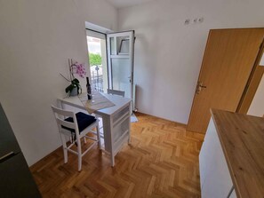 Une petite pièce à aire ouverte, avec un plancher en bois, un bureau blanc avec une chaise, une plante en pot et une porte donnant sur un balcon extérieur.