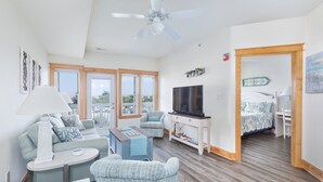 A spacious living room with a ceiling fan, a TV, and a bed in the adjacent room. - C103A- Sweet Sound Memories, Soundfront Nags Head | Sunset Views & Pool (Nags Head)