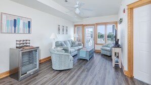 A spacious living room with a grey sofa, a blue armchair, and a wooden coffee table. A glass-fronted refrigerator is on the left, and a ceiling fan is visible. - C103A- Sweet Sound Memories, Soundfront Nags Head | Sunset Views & Pool (Nags Head)
