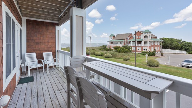 A wooden deck with white chairs, a brick wall, and a view of a large building with a green roof. - C103A- Sweet Sound Memories, Soundfront Nags Head | Sunset Views & Pool (Nags Head)