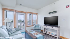 A living room with a sofa, armchair, coffee table, TV stand, and large windows. - C103A- Sweet Sound Memories, Soundfront Nags Head | Sunset Views & Pool (Nags Head)