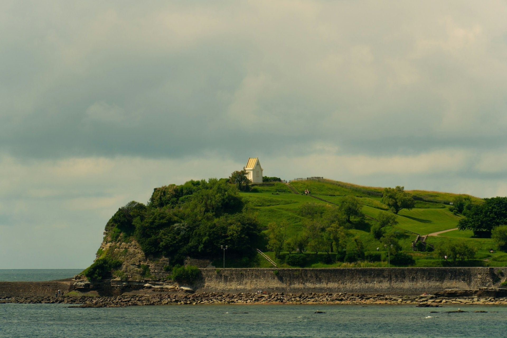 A coastal landscape with a small white building on a grassy hill, a rocky shoreline, and a body of water.