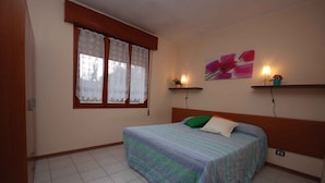 Une chambre à coucher avec un lit, une fenêtre ornée de rideaux en dentelle, une étagère fixée au mur avec une plante et un tableau.