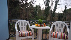 Deux chaises blanches avec des coussins à rayures sur un balcon doté d’une balustrade, une petite table avec une plante et deux boissons, et une vue sur des arbres et un bâtiment au loin.