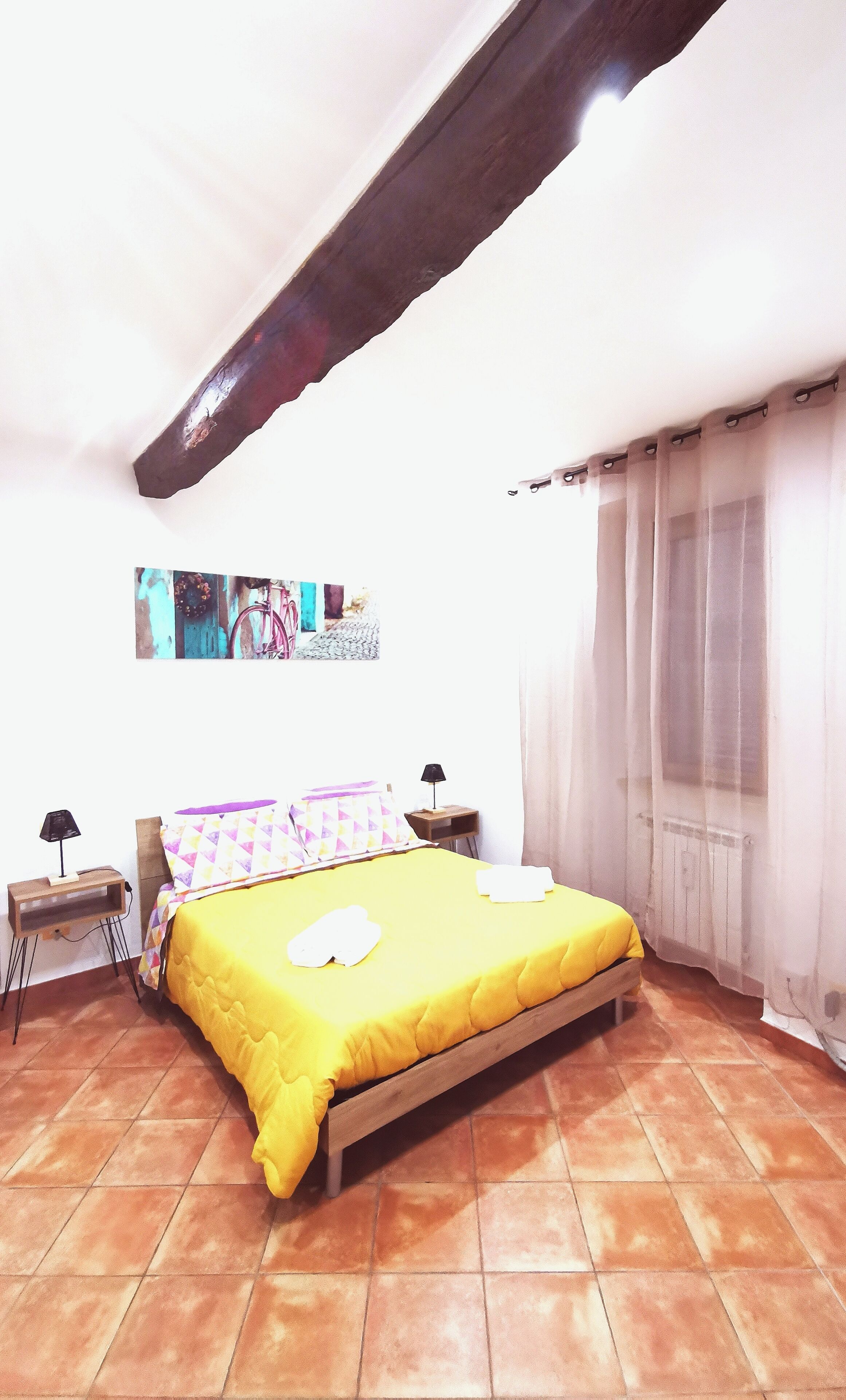 A bedroom with a wooden bed, a small bedside table with a lamp, a wall-mounted shelf, and a window with sheer curtains.