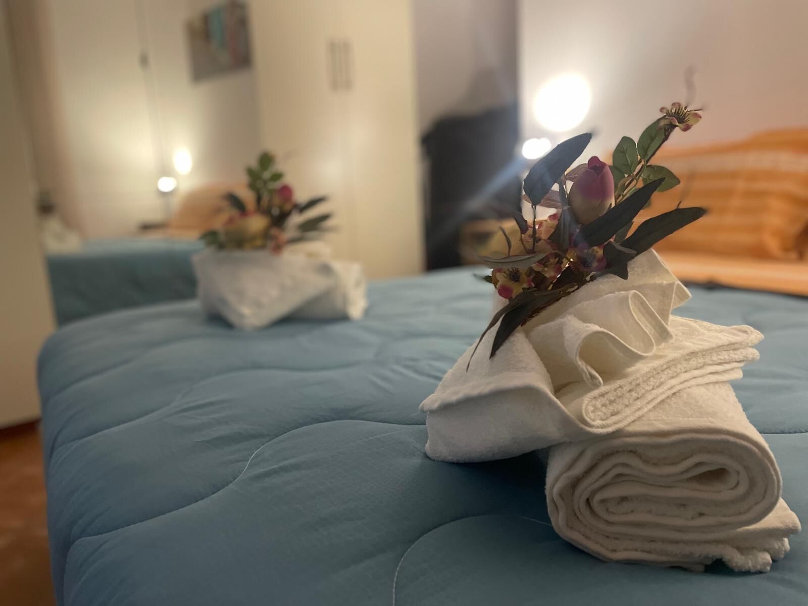 Een hotelkamer met een blauwe spreideken, een keurig opgevouwen witte handdoek op het bed en een kleine potplant op het nachtkastje.