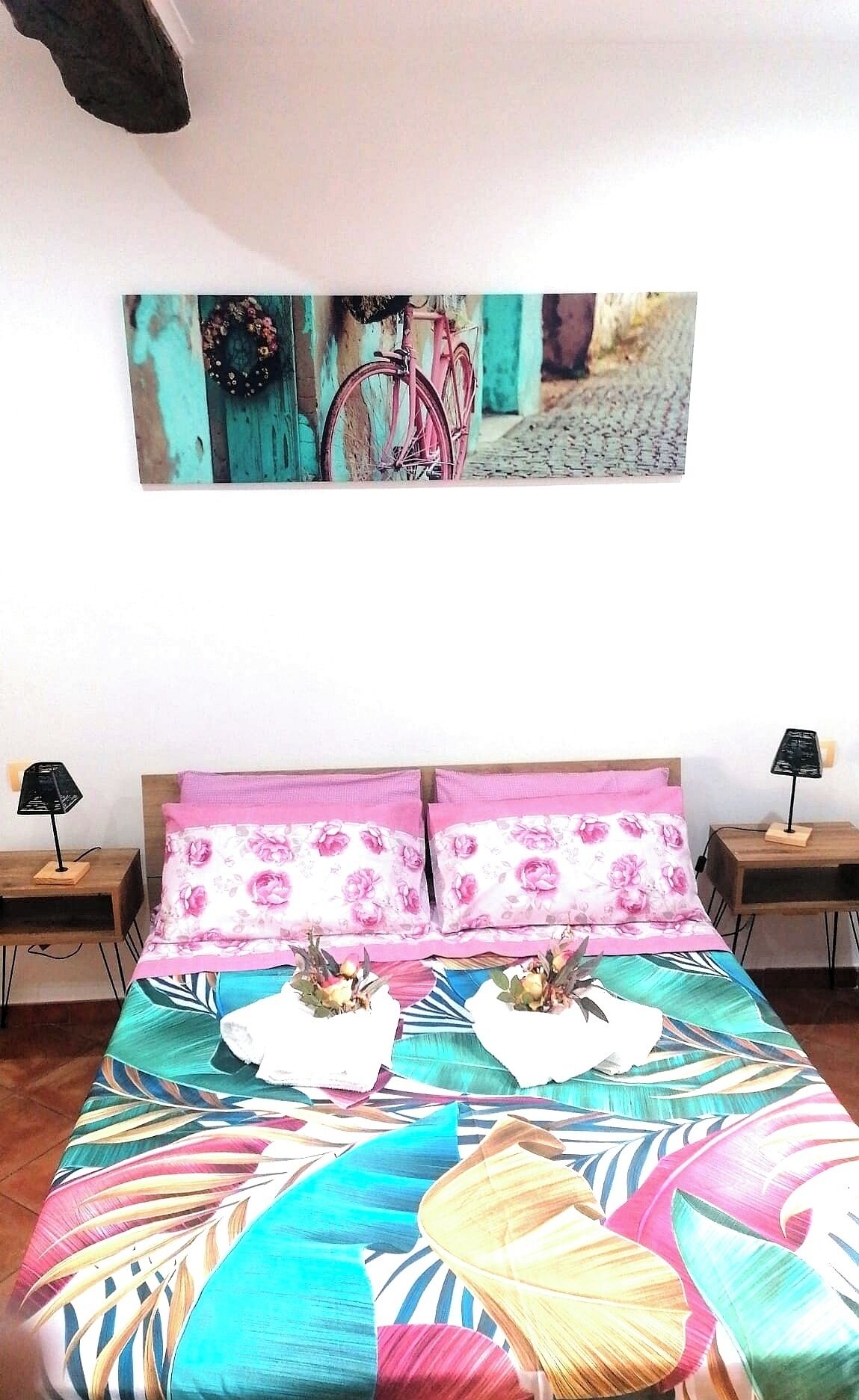 Un lit recouvert d’une couvre-lit aux motifs floraux colorés, deux tables de chevet avec des lampes et un tableau représentant un vélo accroché au mur.