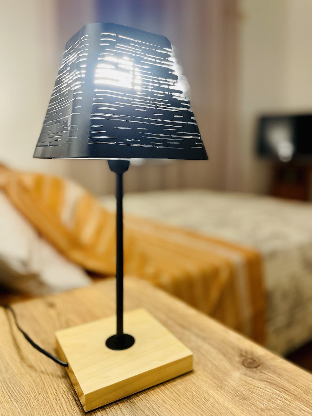 Una lampada moderna con un paralume nero su un tavolo di legno in una stanza con un letto e una coperta gialla.
