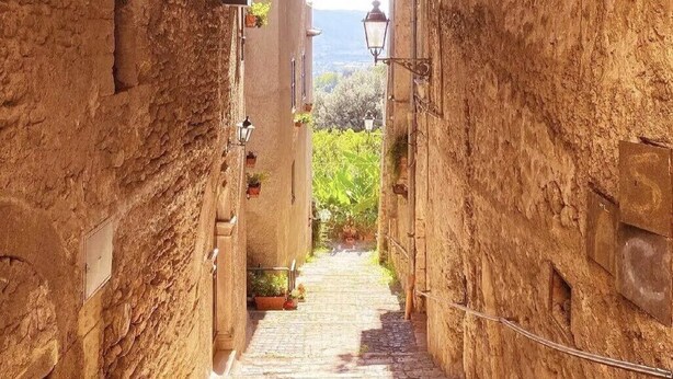 Un étroit sentier de pierre mène à travers une porte en arc, bordé de murs rustiques et illuminé par une lanterne.