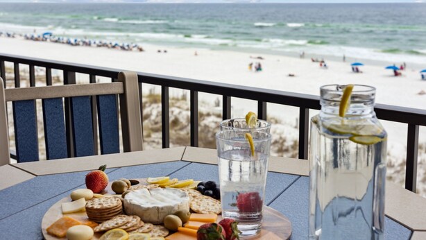 Una mesa con una bandeja de quesos, un vaso de agua y una botella de agua en un balcón con vistas a la playa.