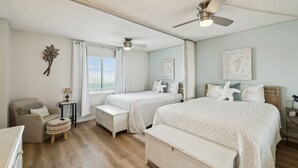 Un dormitorio con dos camas, un ventilador de techo, una ventana con cortinas, una mesita de noche con una lámpara y un adorno de pared.