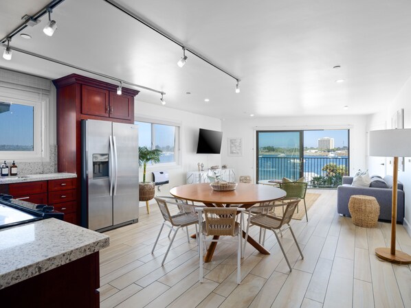 A modern kitchen with a large island, a dining area with wicker chairs, and a balcony with a view of the sea.