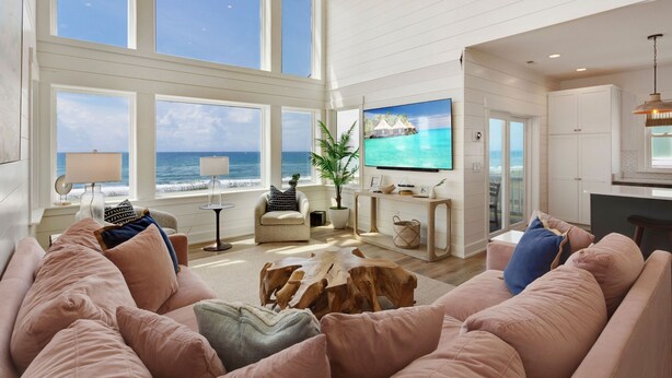 A spacious living room with large windows overlooking the ocean, a comfortable seating area, and a kitchen visible through an open doorway.