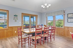 A dining area with a wooden table and chairs, large windows, and a chandelier. - Fore Decs: Private Pool, Tiki Bar & Ocean Views (Corolla)