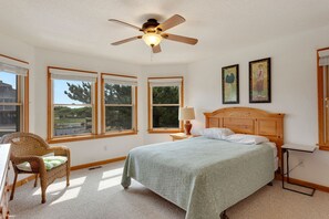 A bedroom with a bed, a chair, a nightstand, a lamp, and two windows. - Fore Decs: Private Pool, Tiki Bar & Ocean Views (Corolla)