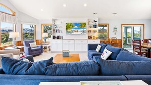 A spacious living room with a large blue sofa, a coffee table, and a bookshelf. There is a dining area with wooden chairs and a table. - Fore Decs: Private Pool, Tiki Bar & Ocean Views (Corolla)
