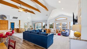 A spacious living area with a large blue sofa, wooden ceiling beams, and a dining area with red chairs. - Fore Decs: Private Pool, Tiki Bar & Ocean Views (Corolla)