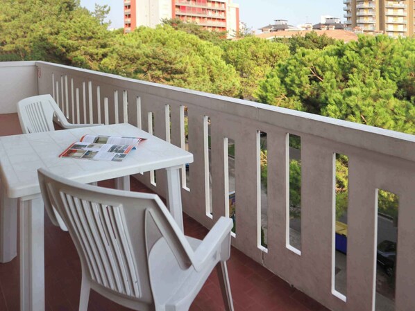 Un balcón con una mesa y sillas blancas, una revista y vistas a árboles y edificios.