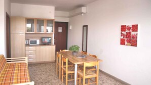 A compact living space with a kitchenette, a dining area with a wooden table and chairs, a sofa, and a potted plant. - Holiday apartment mit Klimaanlage und Balkon (Lignano Sabbiadoro)