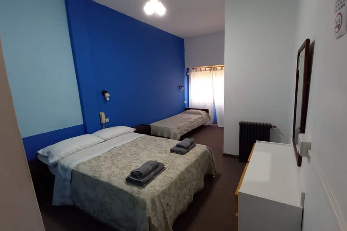 A hotel room with two beds, a desk, a radiator, and a window with curtains.