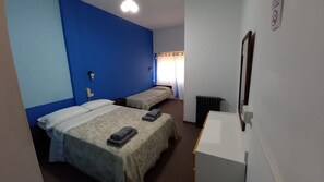 A hotel room with two beds, a desk, a radiator, and a window with curtains.